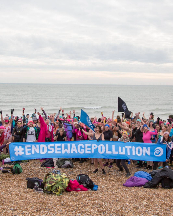 Group of open water swimmers holding a sign saying #EndSewagePollution on the beach with the sea behind